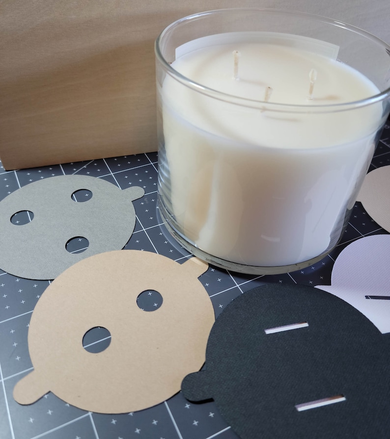 Overhead view of candle dust covers in black, gray, white, and cream on a wood background. Standard and wood wick options.