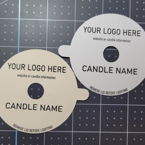 Sample photo of cream and white custom candle dust covers with standard wicks, black text, and example text sitting on top of a measuring grid for size.