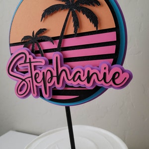 Retro style sunset cake topper in orange, hot pink, purple, neon blue, and black on a white cake with a black cake stick.