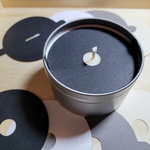 Overview of candle dust covers in a variety of color options, including black, white, cream, gray, and kraft brown. A silver tin candle is featured with a black dust cover in standard wick as an example.