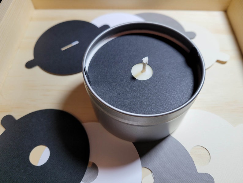 Overview of candle dust covers in a variety of color options, including black, white, cream, gray, and kraft brown. A silver tin candle is featured with a black dust cover in standard wick as an example.