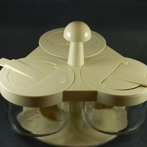 Könnte beinhalten: Ein weißer dreiteiliger Gewürzständer aus Kunststoff mit Glasbehältern und klappbaren Deckeln. Der Ständer hat einen zentralen Knopf in Pilzform.