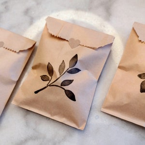 May include: Three brown paper bags with heart-shaped stickers. Each bag has a different black ink stamp design: a cluster of flowers, a leafy branch, and a single flower.