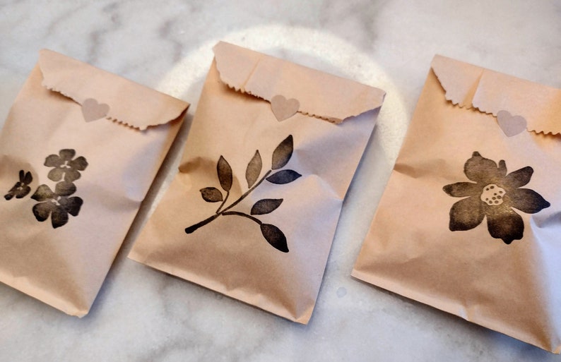 May include: Three brown paper bags with heart-shaped stickers. Each bag has a different black ink stamp design: a cluster of flowers, a leafy branch, and a single flower.