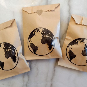 May include: Three brown paper bags with a black globe design and a small heart sticker on each. The bags are on a white marble surface.