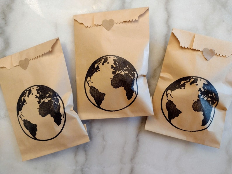 May include: Three brown paper bags with a black globe design and a small heart sticker on each. The bags are on a white marble surface.