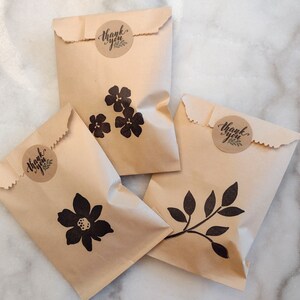May include: Four brown paper bags with thank you stickers and black floral and leaf designs. The bags are on a white marble surface.
