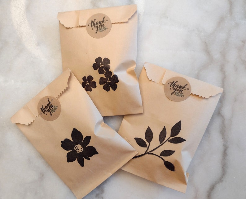May include: Four brown paper bags with thank you stickers and black floral and leaf designs. The bags are on a white marble surface.