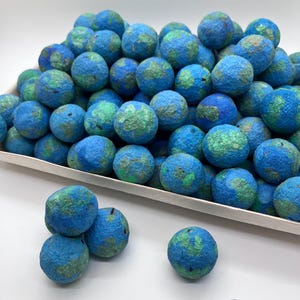 May include: A collection of spherical felted wool balls featuring a mottled pattern of bright blue and green colors. The round items are piled together in a rectangular tray, with several individual spheres resting outside the container.
