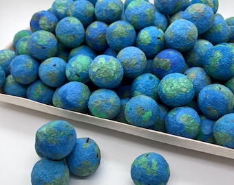May include: A collection of spherical felted wool balls featuring a mottled pattern of bright blue and green colors. The round items are piled together in a rectangular tray, with several individual spheres resting outside the container.