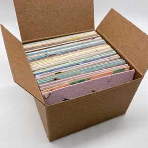 May include: A brown cardboard box filled with a stack of colorful, rectangular cards. The cards have a variety of pastel colors and textures, with visible flecks and patterns. The box is open, revealing the contents inside.