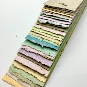 May include: A stack of handmade paper sheets in various pastel colors, including light blue, green, pink, and yellow. The paper has a textured, speckled appearance, with visible flecks of natural fibers.