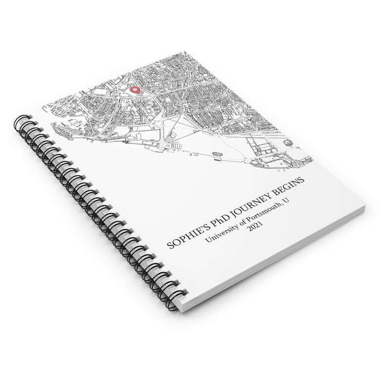 Puede incluir: Una libreta de espiral blanca con un mapa en blanco y negro de una ciudad en la portada. El mapa tiene un alfiler rojo que marca una ubicaci&oacute;n. El texto "SOPHIE'S PHD JOURNEY BEGINS" est&aacute; impreso en negro en la portada, junto con "University of Portsmouth, UK" y "2021".