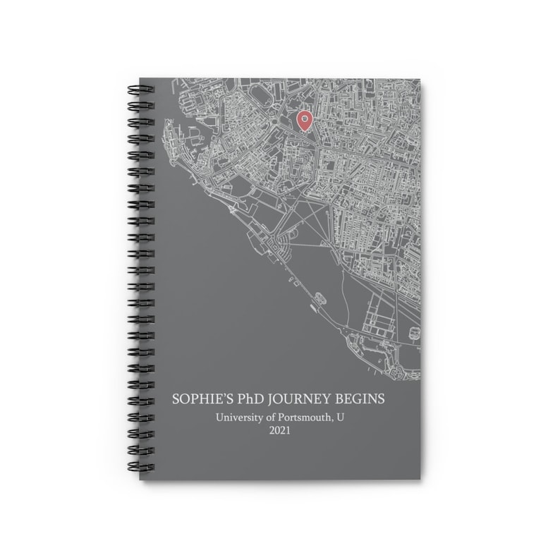 Puede incluir: Un cuaderno de espiral gris con un mapa de una ciudad y un alfiler rojo que marca una ubicaci&oacute;n. El texto en la portada dice "Sophie's PhD Journey Begins, University of Portsmouth, 2021."