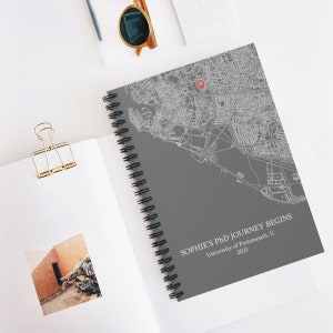 Puede incluir: Una libreta espiral gris con un mapa de una ciudad y el texto "Sophie's PhD Journey Begins University of Portsmouth, U 2021" impreso en la portada.