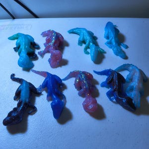 May include: An assortment of eight small, lizard-shaped figurines in various colors. The figurines are in shades of blue, pink, and purple, some with glitter accents. They are arranged on a white surface.