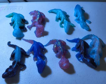 May include: An assortment of eight small, lizard-shaped figurines in various colors. The figurines are in shades of blue, pink, and purple, some with glitter accents. They are arranged on a white surface.
