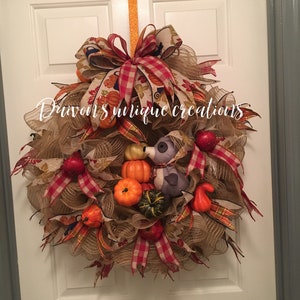 Op de afbeelding: Een herfstkrans met een jute basis, versierd met pompoenen, kalebassen en rode appels. De krans heeft een grote strik met herfstkleurige linten en de tekst "Duvon's unique creations". Een oranje lint wordt gebruikt om op te hangen.