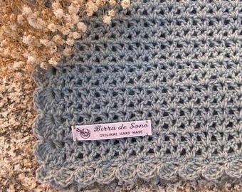 May include: A light blue crocheted blanket with a scalloped edge and a label that reads "Birra de Sono Original Hand Made".
