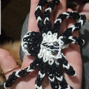 May include: A handmade spider crafted from black and white rubber bands. The spider has eight legs, a round body, and iridescent bead eyes. The spider is a unique, decorative item.