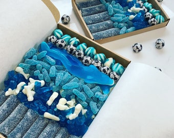 May include: A blue and white gift box filled with blue and white candy. The candy is arranged in a pattern that resembles a wave. The box is open and the candy is visible.