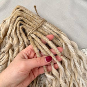 Pu&ograve; includere: Un fascio di dreadlock sintetici biondi con un aspetto ondulato e strutturato. Le singole ciocche sono legate insieme nella parte superiore con uno spago sottile color naturale. Le extension presentano un colore biondo sabbia chiaro con radici marrone scuro nel punto di attacco, creando un effetto multitonale lungo tutta la lunghezza dei capelli ondulati.