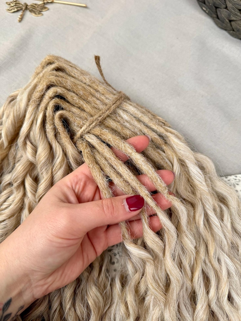 Pu&ograve; includere: Un fascio di dreadlock sintetici biondi con un aspetto ondulato e strutturato. Le singole ciocche sono legate insieme nella parte superiore con uno spago sottile color naturale. Le extension presentano un colore biondo sabbia chiaro con radici marrone scuro nel punto di attacco, creando un effetto multitonale lungo tutta la lunghezza dei capelli ondulati.