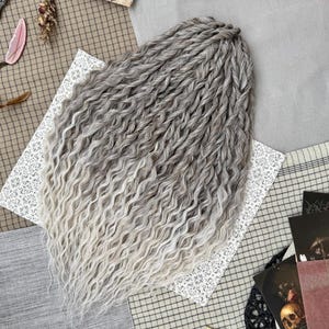 May include: A set of synthetic hair extensions featuring a gradient colour transition from dark grey at the roots to a light platinum blonde at the ends. The hair is styled in tight, wavy twists that create a voluminous, textured appearance.