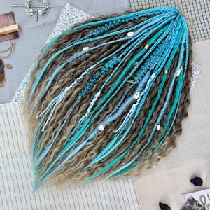 Puede incluir: Un conjunto de rastas hechas a mano en tonos marrón, azul y blanco. Las rastas están adornadas con pequeños anillos dorados, conchas blancas y detalles florales azules. Los extremos de las rastas tienen una textura ondulada.