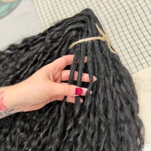 May include: A bundle of black synthetic hair extensions featuring a wavy, textured finish. The individual strands are gathered together and secured at the top with a piece of natural-colored twine, creating a uniform and organized hair piece.