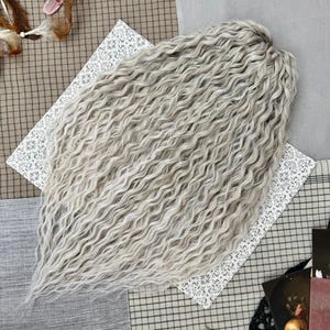 May include: A bundle of wavy, light gray hair extensions. The hair is styled in loose, flowing waves. The extensions are laid on a white patterned fabric, with a checkered and solid gray background.