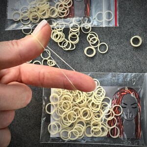 May include: A collection of small, translucent, off-white elastic hair bands packaged in clear plastic bags. Each bag features a graphic illustration of a face with long, styled hair. One elastic band is stretched thin between two fingers.