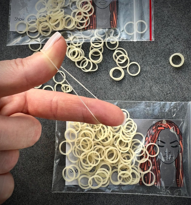 May include: A collection of small, translucent, off-white elastic hair bands packaged in clear plastic bags. Each bag features a graphic illustration of a face with long, styled hair. One elastic band is stretched thin between two fingers.
