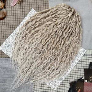 May include: A set of light blonde hair extensions featuring a wavy, crimped texture. The strands are arranged in a dense, layered cluster, showcasing a consistent pale, ash-toned color throughout the length of the wavy synthetic hair fibers.