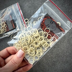 May include: A collection of small, off-white circular elastic bands packaged in a clear plastic resealable bag. Inside the bag is a card featuring a graphic illustration of a person with long, reddish-brown braided hair and closed eyes.