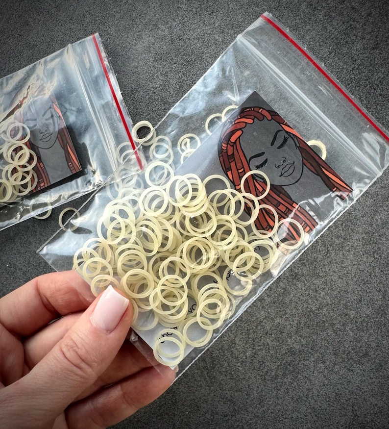 May include: A collection of small, off-white circular elastic bands packaged in a clear plastic resealable bag. Inside the bag is a card featuring a graphic illustration of a person with long, reddish-brown braided hair and closed eyes.