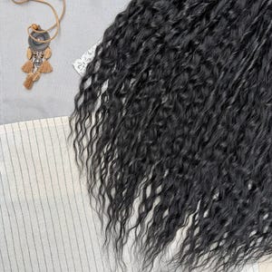 May include: A bundle of long, black, wavy hair extensions with a crimped texture. The hair is arranged in a dense, flowing mass, showcasing a consistent dark color and a defined, wavy pattern throughout the length of the strands.
