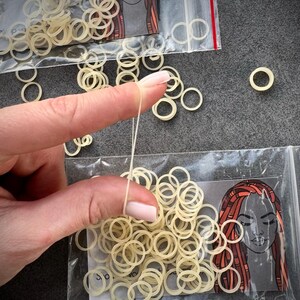 May include: A collection of small, translucent, off-white elastic hair bands packaged in clear plastic bags. Each bag features a printed illustration of a person with long, reddish-brown dreadlocks. The bands are circular, flexible, and thin.