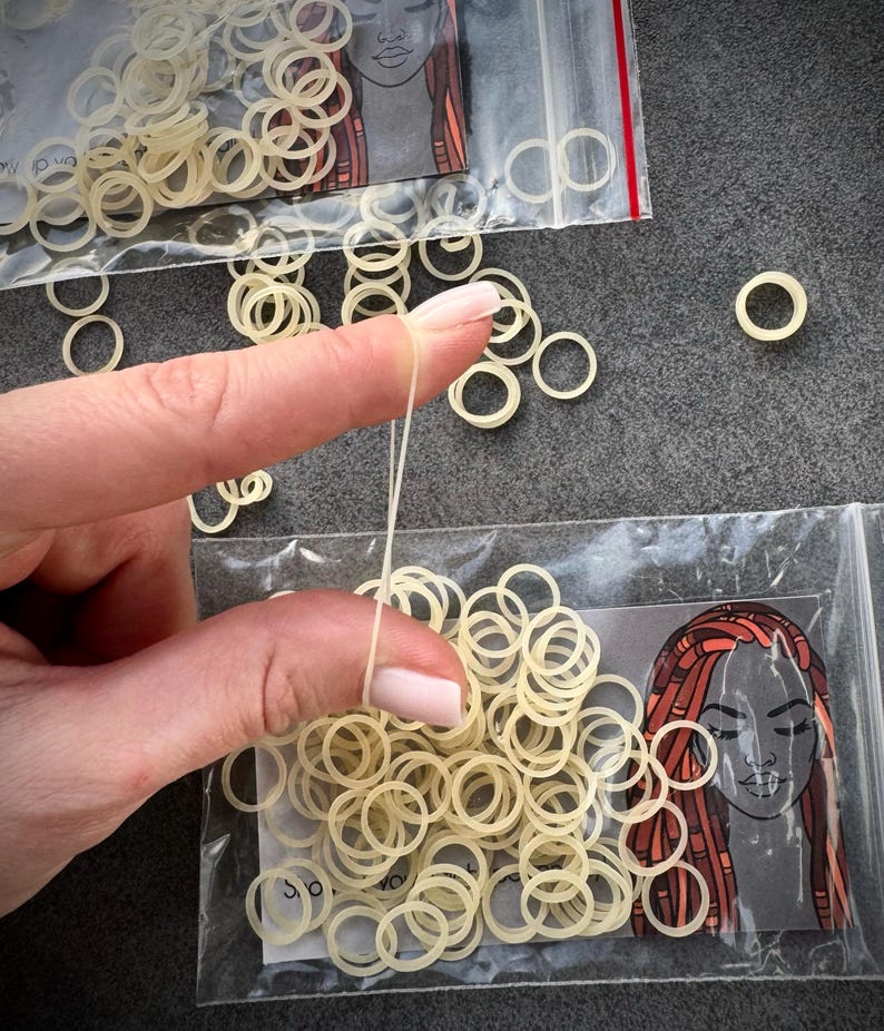 May include: A collection of small, translucent, off-white elastic hair bands packaged in clear plastic bags. Each bag features a printed illustration of a person with long, reddish-brown dreadlocks. The bands are circular, flexible, and thin.