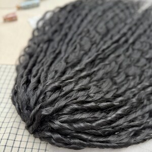 May include: A bundle of long, dark charcoal-black hair extensions featuring a tight, twisted, and wavy texture. The individual strands are densely packed together, creating a voluminous and uniform appearance with a consistent dark color throughout.