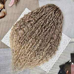 May include: A set of synthetic hair extensions featuring a wavy, crimped texture. The hair transitions from a medium ash brown at the top to a lighter, sandy blonde shade at the ends, creating a multi-tonal, highlighted appearance throughout.