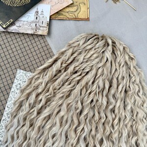 Pu&ograve; includere: Un set di extension per capelli sintetici biondo chiaro caratterizzato da una consistenza stretta, ondulata e ritorta. Le ciocche sono disposte in un fascio denso e uniforme, mostrando una tavolozza di colori biondi con sottili variazioni.