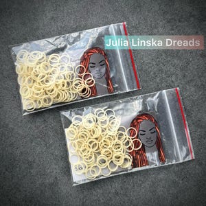 May include: Two clear plastic resealable bags containing small, off-white elastic hair bands. Each bag features a printed card insert showing an illustration of a person with long, reddish-brown dreadlocks and the text Julia Linska Dreads.