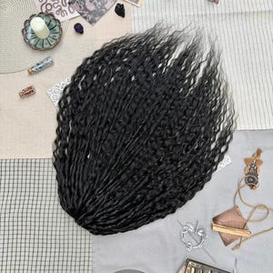 May include: A bundle of long, black, wavy hair extensions with a crimped texture. The hair is arranged in a fan shape, showcasing the dark color and the consistent, tight wave pattern throughout the length of the individual strands.