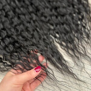 May include: A bundle of long, black hair extensions with a distinct wavy texture. The strands are fine and dark, showcasing a natural-looking curl pattern that flows downward. The hair appears soft and lightweight, with individual strands visible.