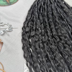 May include: A set of long, dark charcoal-colored synthetic hair extensions featuring a tight, wavy, and crimped texture. The strands are bundled together, showcasing a consistent dark tone and a wavy pattern throughout the length of the hair pieces.