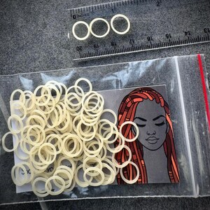 May include: A clear plastic bag containing numerous small, off-white elastic hair bands. A rectangular card inside the bag features an illustration of a person with long, reddish-brown locs. A clear plastic ruler shows the bands are 0.25 inches wide.