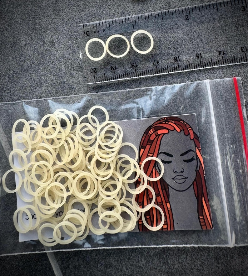 May include: A clear plastic bag containing numerous small, off-white elastic hair bands. A rectangular card inside the bag features an illustration of a person with long, reddish-brown locs. A clear plastic ruler shows the bands are 0.25 inches wide.