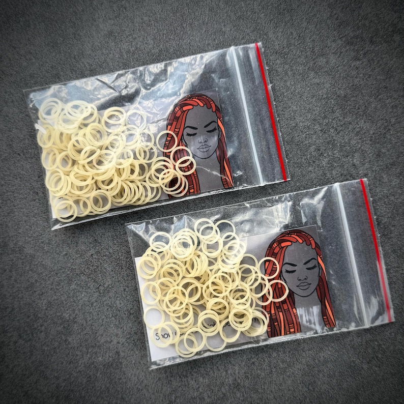 May include: Two clear plastic resealable bags containing small, off-white elastic hair bands. Each bag features a printed graphic of a person with long, reddish-brown hair and dark skin tones, positioned next to the pile of elastic bands.