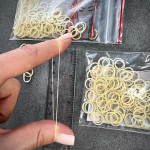 May include: Two clear plastic bags filled with small, off-white elastic hair bands. One bag features a graphic illustration of a person with braided hair. A clear plastic ruler shows one elastic band stretched to approximately 2.5 inches in length.